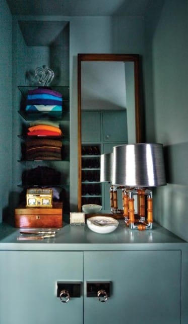 Blue closet interior with glass shelves holding folded colorful towels and sweaters, a wooden jewelry box, and a silver lamp with bamboo accents on a gray countertop.