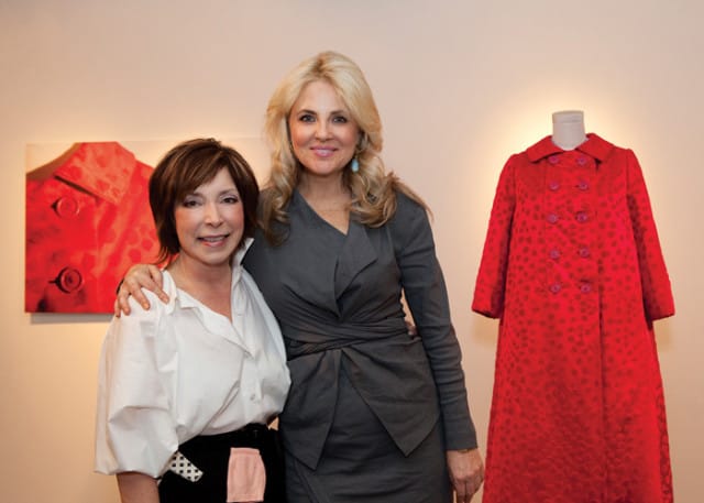 Two women stand in front of a red coat artwork and a red coat on a mannequin. The woman on the left wears a white blouse and black skirt. The woman on the right wears a gray wrap dress.