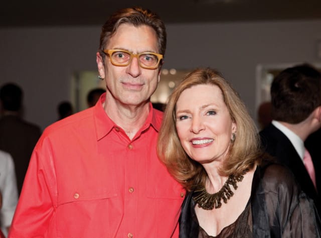 Man with yellow glasses and a coral shirt stands next to a woman wearing a dark, sheer top and a statement necklace.