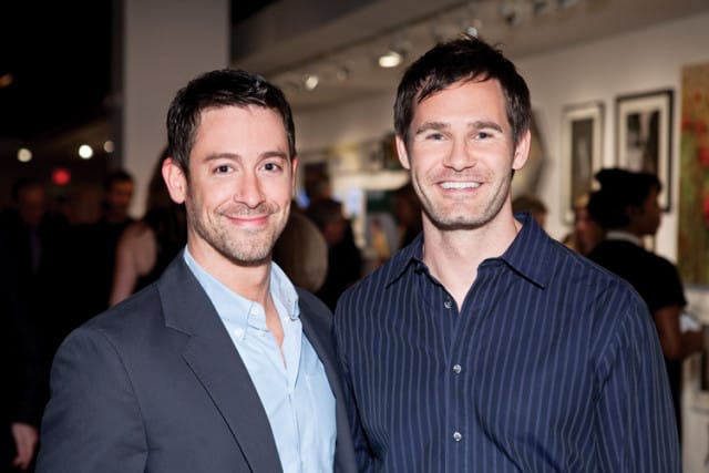 Two men smiling at the camera, one in a dark suit jacket and light blue shirt, the other in a dark pinstripe shirt. They are at an event with other people and artwork in the background.