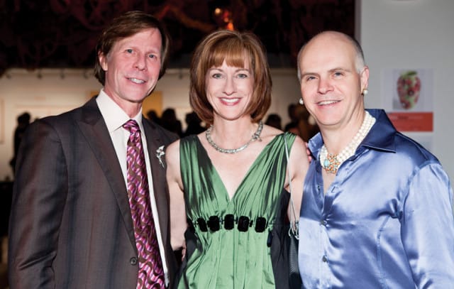 Three people posing for a photo at an event. A man in a suit and patterned tie stands on the left. A woman in a green dress and necklace is in the middle. A person in a blue satin shirt and pearl necklaces stands on the right.