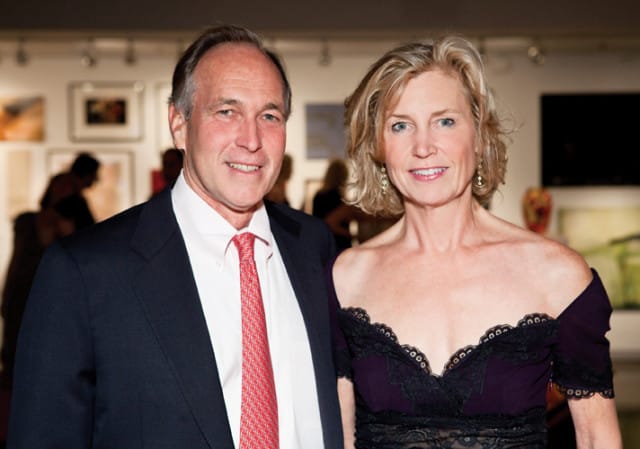 Man in a navy suit and red patterned tie stands next to a woman in a purple off-the-shoulder dress with black lace trim. They are at an event with artwork on the walls behind them.
