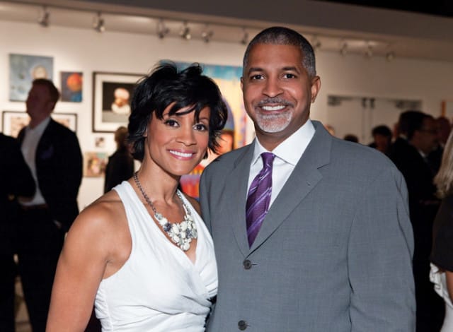 A woman in a white dress and a man in a gray suit with a purple tie smile at an event with artwork on the wall behind them.