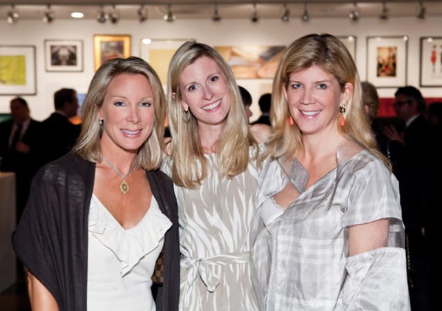 Three women with blonde hair smile for the camera at an event. The woman on the left wears a white top and dark shawl, the middle woman wears a patterned dress, and the woman on the right wears a light-colored plaid outfit.