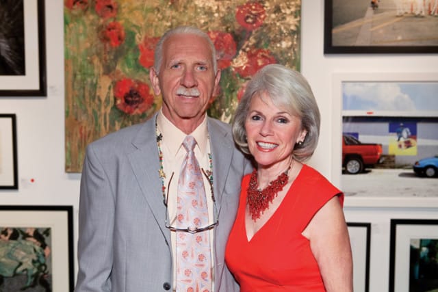 Couple posing in front of artwork at an event. The man wears a light gray suit, peach shirt, and patterned tie. The woman wears an orange dress and a red beaded necklace.