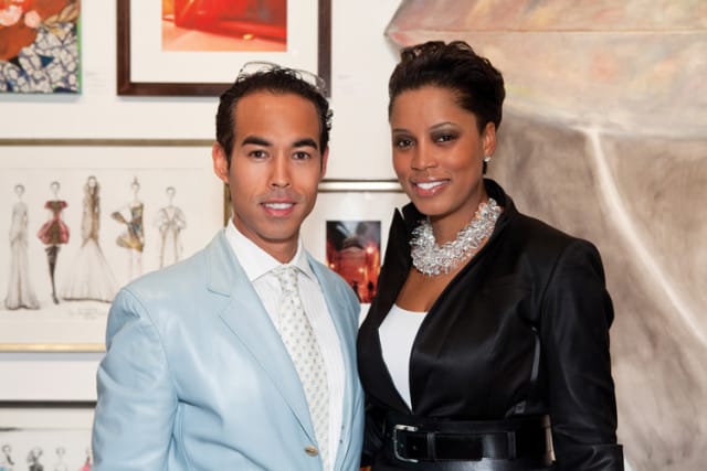 Man in a light blue suit and woman in a black jacket with a statement necklace pose in front of artwork.