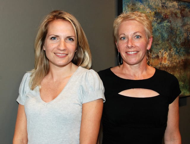 Two women posing for a portrait, with a colorful abstract painting in the background. The woman on the left has blonde hair and is wearing a light grey v-neck shirt. The woman on the right has short blonde hair and is wearing a black shirt with a cutout detail.