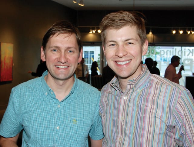Two men smiling at the camera. The man on the left wears a teal checkered button-down shirt. The man on the right wears a striped button-down shirt with blue, pink, and yellow stripes.