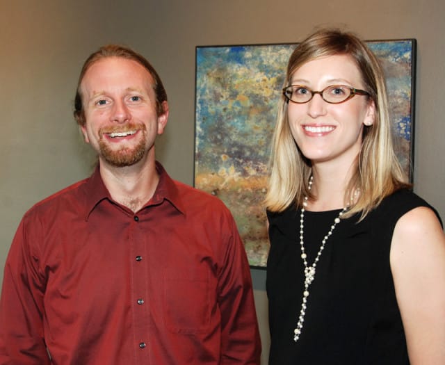 Man with beard wearing a red collared shirt and woman wearing a black dress and pearl necklace, standing in front of abstract art.