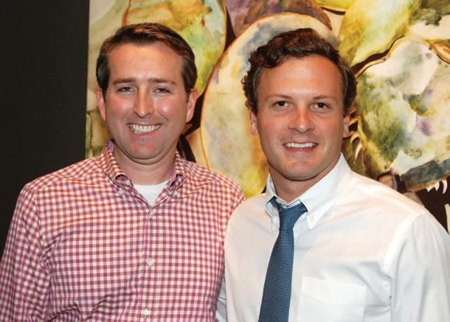 Two men smiling in front of a colorful abstract painting. The man on the left wears a pink and white gingham shirt, and the man on the right wears a white button-down shirt and a blue tie.