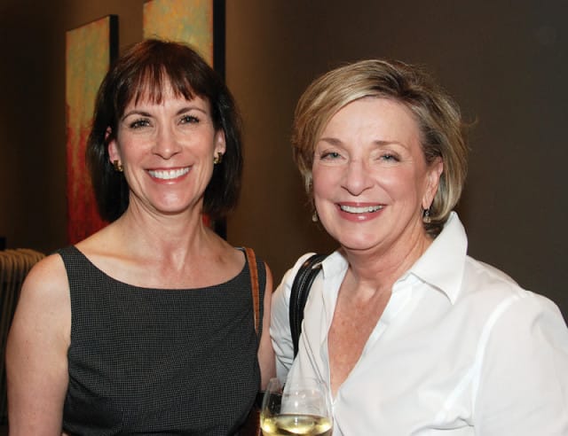 Two women smiling at the camera. The woman on the left wears a dark patterned sleeveless dress, and the woman on the right wears a white button-down shirt.
