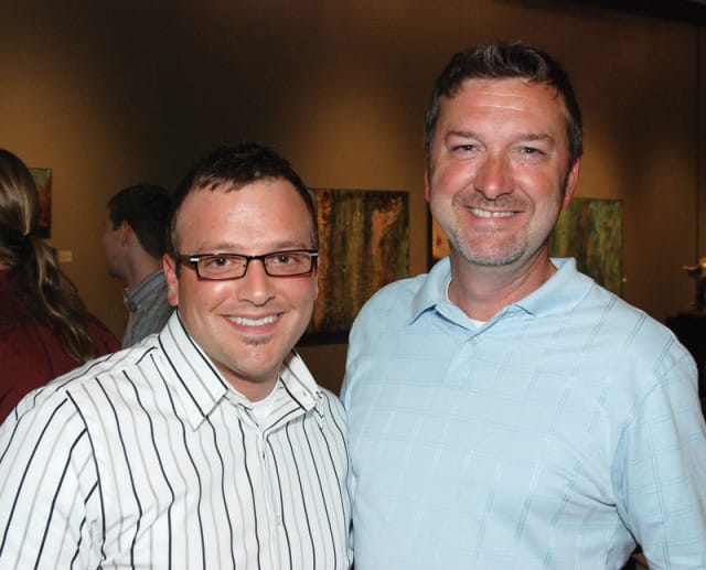 Two men smiling at the camera. The man on the left wears glasses and a striped shirt. The man on the right wears a light blue polo shirt.