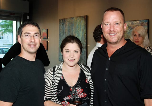 Three people posing for a photo at an art gallery event. The man on the left wears glasses and a black t-shirt. The woman in the center wears a black and white striped cardigan over a floral dress. The man on the right wears a black button-down shirt.