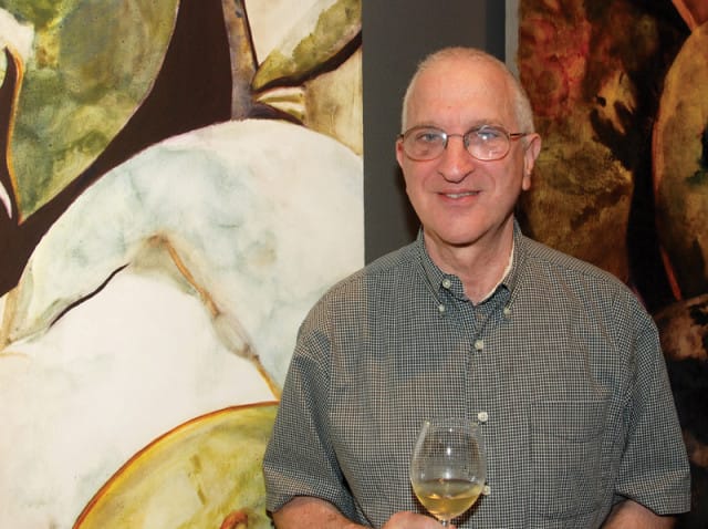 Man with glasses and a checkered shirt holds a glass of white wine in front of abstract paintings.