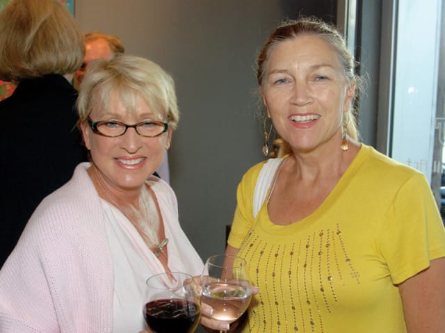Two women smiling and holding wine glasses. The woman on the left wears glasses and a pink cardigan over a white top. The woman on the right wears a yellow shirt with beaded embellishments.
