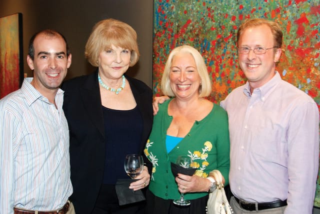 Four people smile for a photo in front of a colorful abstract painting. A man in a striped shirt stands to the left, next to a woman in a black blazer and turquoise necklace. A woman in a green cardigan and a man in a light purple shirt stand to the right.