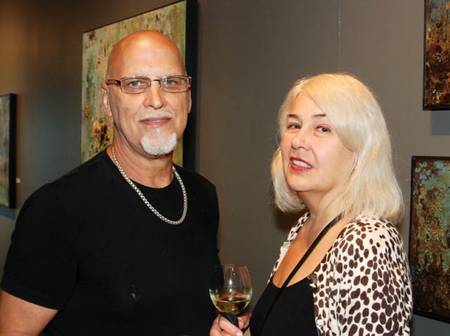 Man with goatee and glasses wearing a black t-shirt and silver chain, standing next to a woman with blonde hair wearing a leopard print cardigan and black top. Both are holding wine glasses.