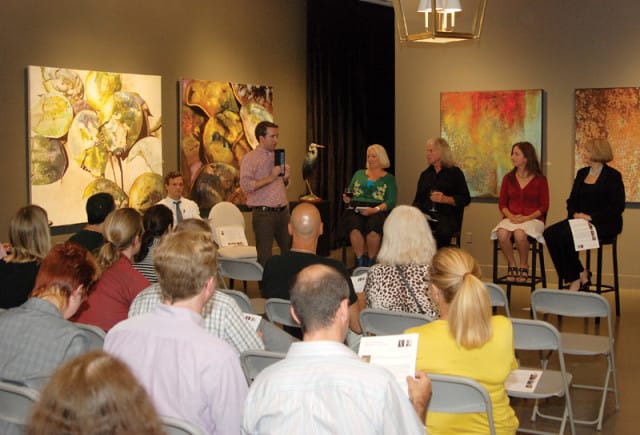 A man stands holding a tablet in front of a seated audience, with artwork on the wall behind him and a panel of four women seated on chairs.