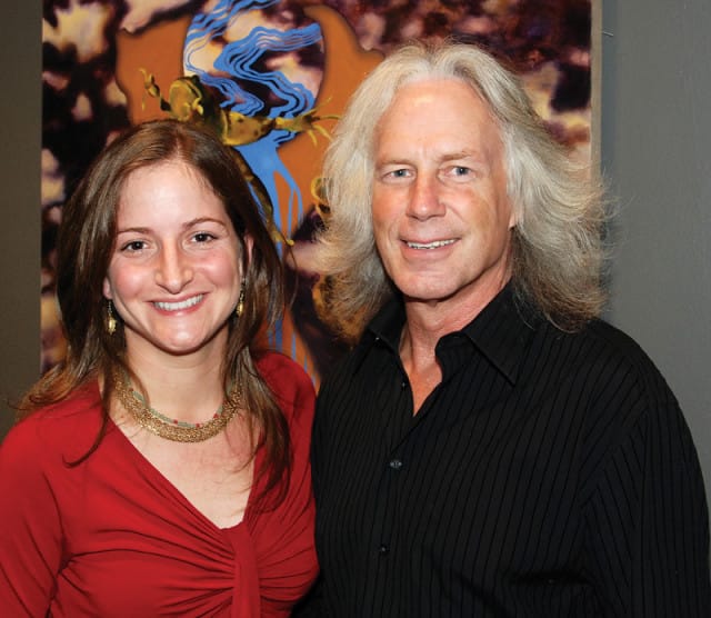 A woman in a red top and a man with long gray hair in a black striped shirt smile at the camera in front of abstract art.