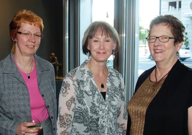 Three women smiling at the camera. The woman on the left wears a gray plaid jacket over a pink top and glasses. The woman in the middle wears a black and white patterned blouse and earrings. The woman on the right wears glasses and a black top over a gold textured shirt.