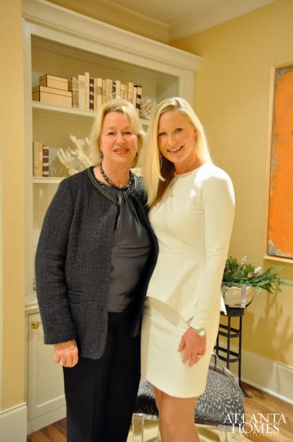Two women posing in front of a bookshelf. The woman on the left wears a dark textured jacket over a gray top and black pants. The woman on the right wears a white peplum dress.