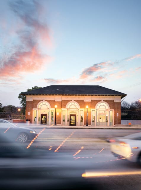 Brick train station with arched windows and doors, illuminated by streetlights at dusk, with blurred cars in the foreground.