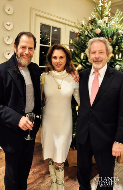 Three people pose in front of a Christmas tree. The man on the left holds a champagne flute. The woman in the center wears a white dress and tall boots. The man on the right wears a suit and pink tie.