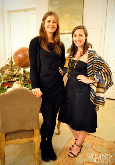 Two women smiling, one in a black dress and cardigan, the other in a black dress and striped shawl. A globe and decorative display are behind them.
