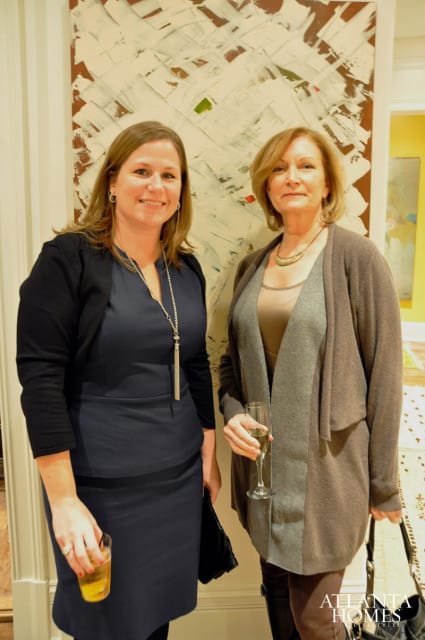 Dee Murphy and Betsy McBride stand in front of abstract art. Murphy wears a navy dress and black cardigan, holding a drink. McBride wears a brown cardigan over a taupe top, holding a champagne flute.