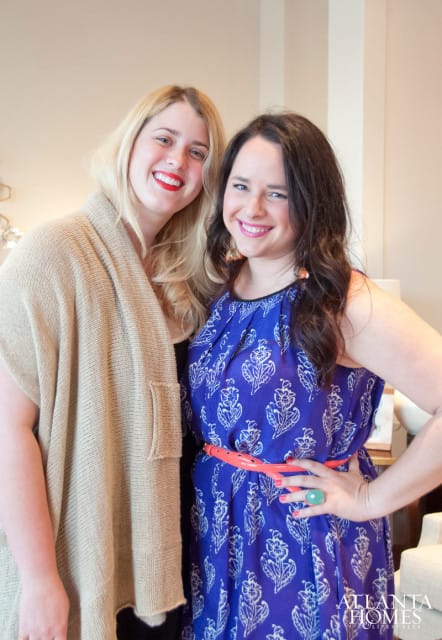 Two women smiling at the camera. The woman on the left has blonde hair and wears a tan knit cardigan. The woman on the right has dark hair and wears a blue dress with a white floral pattern and a coral belt.