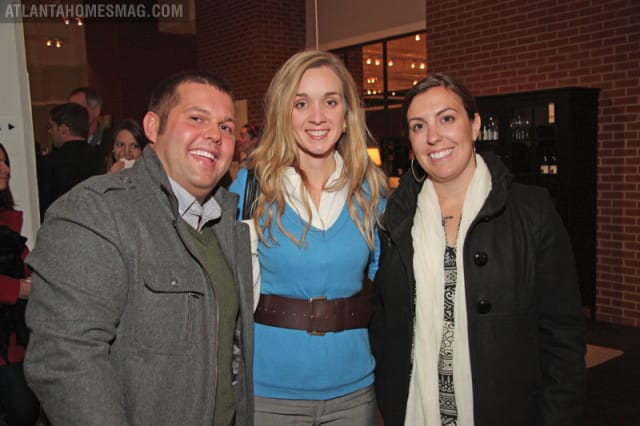 Three smiling people pose for a photo. The man on the left wears a grey coat over a green sweater. The woman in the middle wears a blue sweater and a brown belt. The woman on the right wears a black coat and a white scarf.