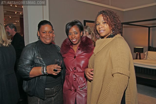 Three women posing for a photo. The woman on the left wears a black leather jacket and holds a drink. The woman in the middle wears a burgundy leather coat with a fur collar. The woman on the right wears a tan knit poncho.