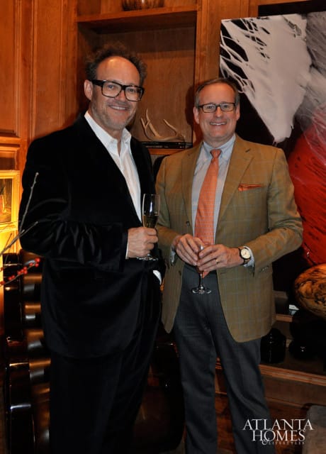 Two men in suits holding champagne glasses in a room with wood paneling and abstract art.