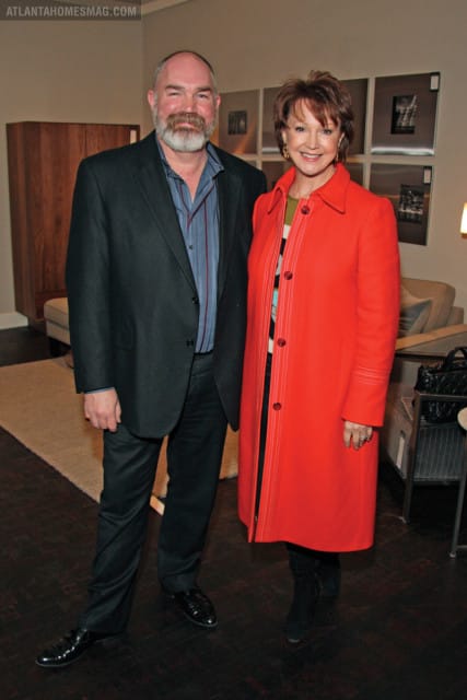 Man in dark suit and striped shirt with a beard and woman in a bright orange coat with a patterned dress stand together indoors.