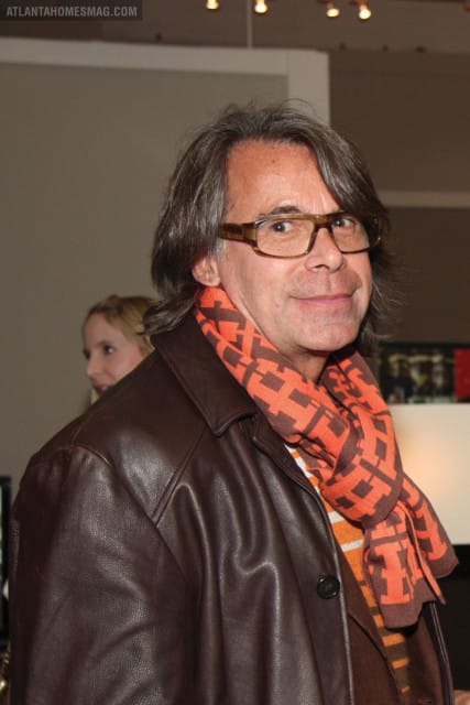 Man with long brown hair, glasses, brown leather jacket, and an orange and brown patterned scarf. A woman is blurred in the background.