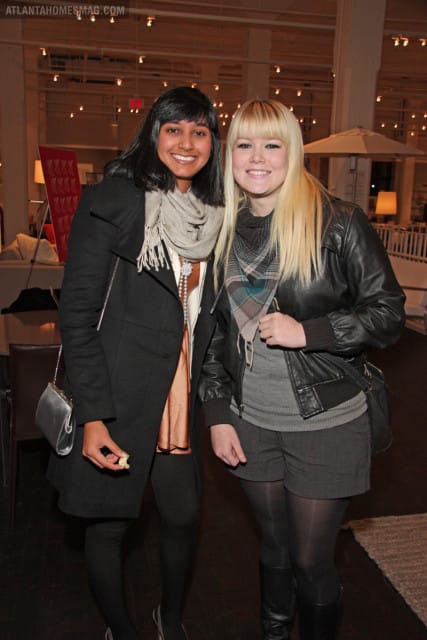 Two smiling women stand together indoors, one with dark hair wearing a black coat and scarf, the other with blonde hair wearing a leather jacket and scarf.