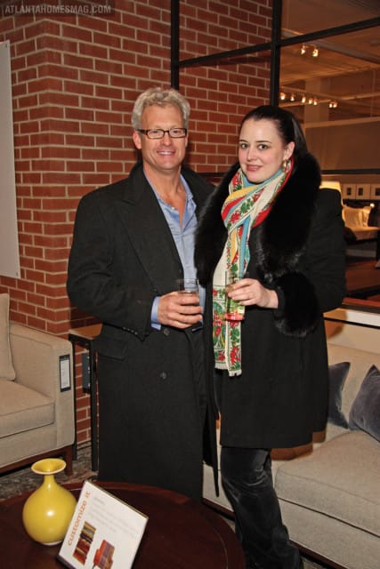 Man in glasses and dark coat smiles next to a woman wearing a black coat with fur trim and a colorful scarf, both holding drinks.