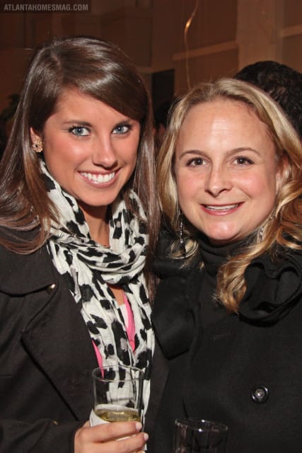 Two smiling women pose for a photo, one with dark hair wearing a black coat and a black and white leopard print scarf, the other with blonde hair wearing a black coat.