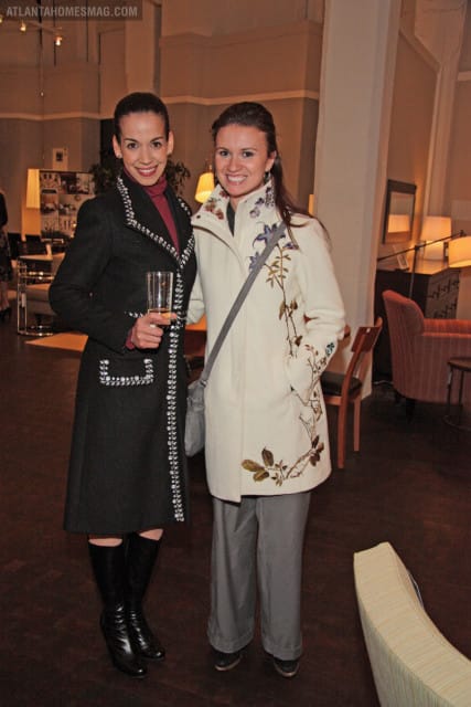 Two women pose for a photo, one in a black coat with jeweled trim and a maroon turtleneck, the other in a cream coat with floral embroidery and gray pants.