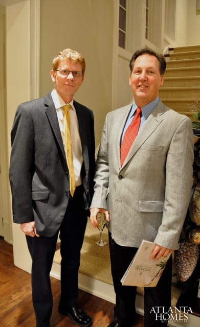 Two men in suits stand near a staircase. The man on the left wears a dark suit with a yellow tie. The man on the right wears a light gray suit with a red tie and holds a magazine.