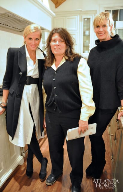 Three women in a kitchen; one on the left wears a black coat over a white shirt and black belt, the middle woman wears a black vest over a yellow shirt and black pants, and the woman on the right wears a black turtleneck.