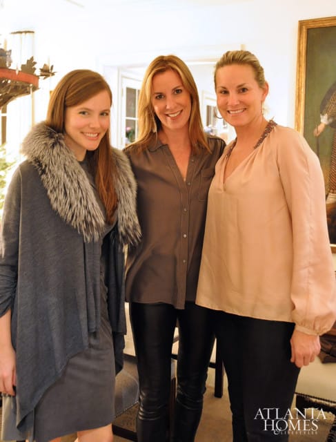 Three women smiling, two wearing leather pants, in a well-lit room with a painting on the wall.