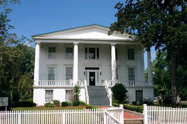 White neoclassical mansion with a portico supported by large columns, a wide staircase leading to the entrance, and a white picket fence in the foreground.