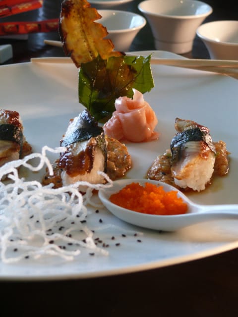 Plate of eel sushi with pickled ginger, orange tobiko roe, and crispy noodles, garnished with a fried plantain leaf