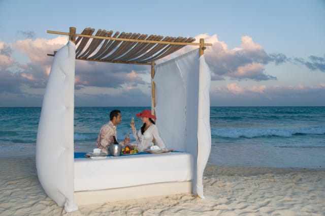 Couple enjoying a romantic dinner on a beach under a bamboo and white fabric canopy, with champagne and fruit platter on a low table.
