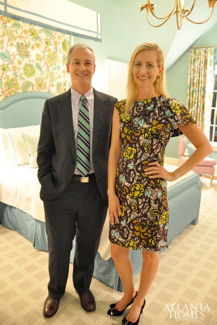 A man in a suit and a woman in a floral dress stand in a room with a light blue bed and patterned wallpaper.