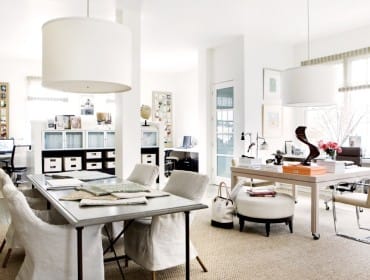 Bright office space with a large metal table and linen-covered chairs in the foreground, and two desks with computers and storage cabinets in the background.