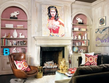 Living room with a Wonder Woman painting above a fireplace, built-in bookshelves with decorative items, a brown leather armchair with a patterned pillow, and a dark sofa.