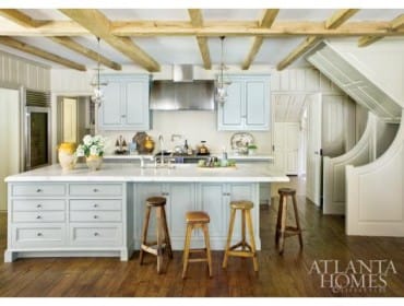 Light blue kitchen with marble island, stainless steel appliances, wooden beams on ceiling, and wooden floors.