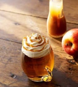 Glass of apple cider topped with whipped cream and caramel drizzle, next to a red apple on a wooden surface.